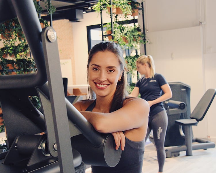 Een vrouwelijk lid van The Gym Society dat met een grote glimlach op een toestel leunt